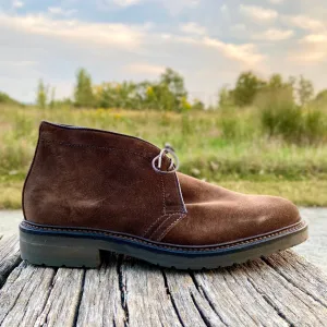 The Alden Madison 1273S - Chukka Boot in Brown Suede, featuring dark stitching and a thick lug rubber sole, rests on weathered wood with a grassy landscape and cloudy sky blurred in the background.