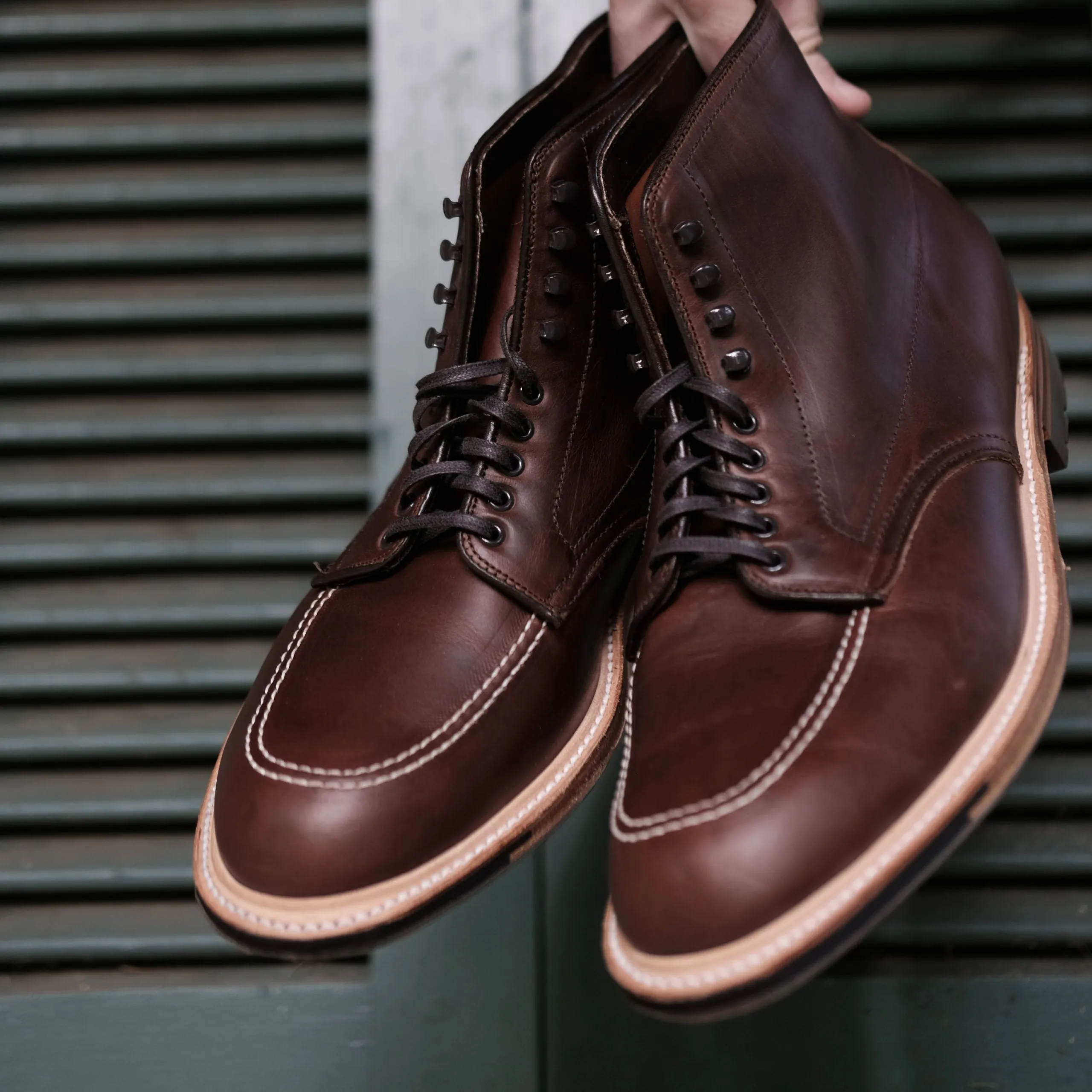 A hand holds a pair of Alden Madisons 403C - Indy Boot in Chromexcel leather, featuring white stitching, the Truebalance Last, and a rugged Commando Sole against a green slatted surface.