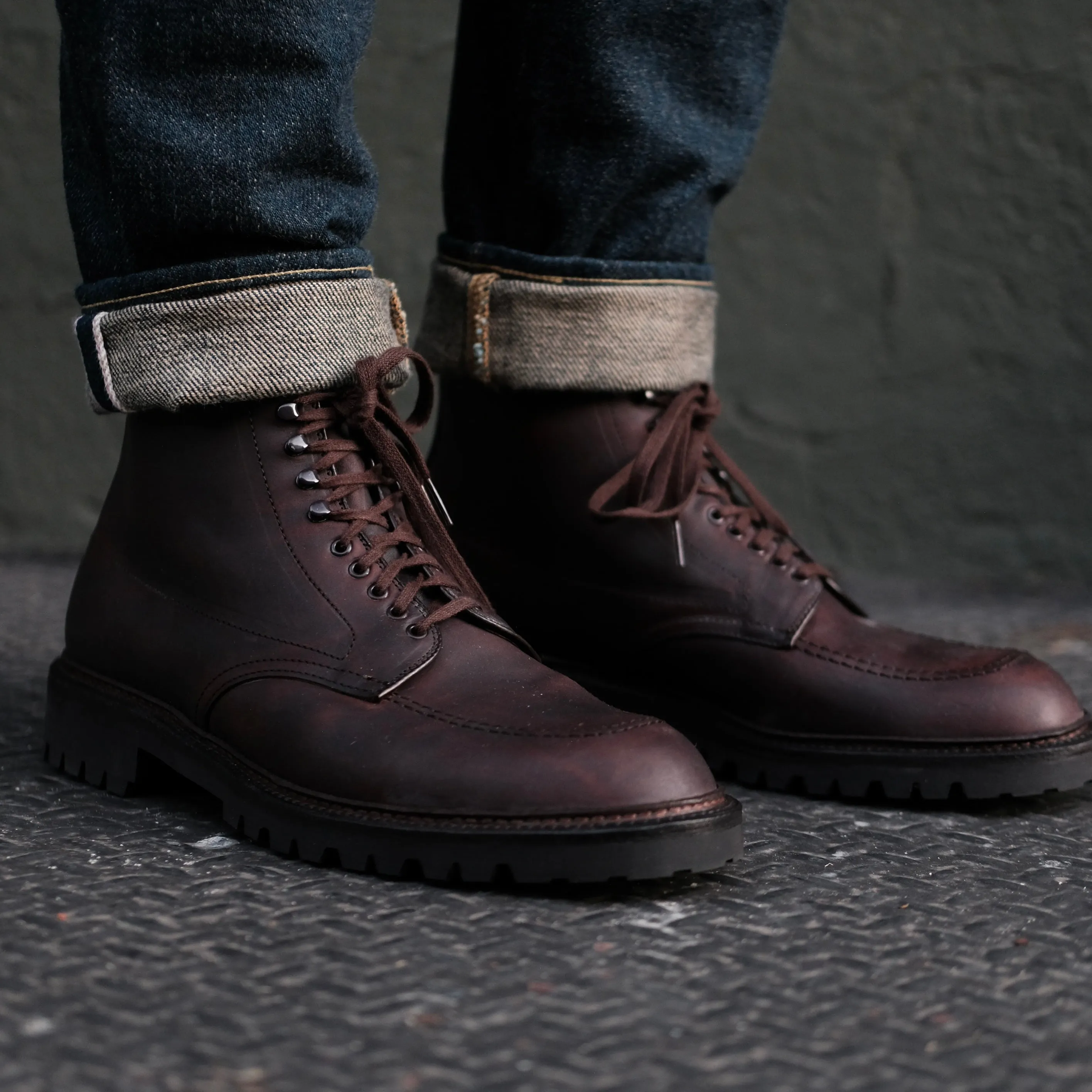 A person is wearing Alden Madisons 404 - Indy Boot in Kudu (Oiled CXL), polished dark brown lace-up boots with a durable Vibram Lug Outsole. Theyre paired with cuffed dark blue jeans and resting on a textured metal surface against a muted green wall.