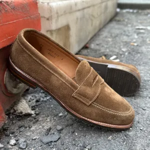 The Alden Madison 6221L - Unlined Penny Mocc in Snuff Suede, rests on gravel near a red and white concrete block. Featuring wooden heels, one shoe leans slightly on the other, creating a casual yet stylish scene.