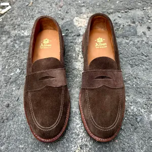 The 6224L Unlined Penny Mocc in Brown Suede by Alden Madison showcases a mocc toe design with stitching details on a rough gray surface. The insoles feature A. Albert in gold, paired with durable rubber soles for enhanced comfort and grip.