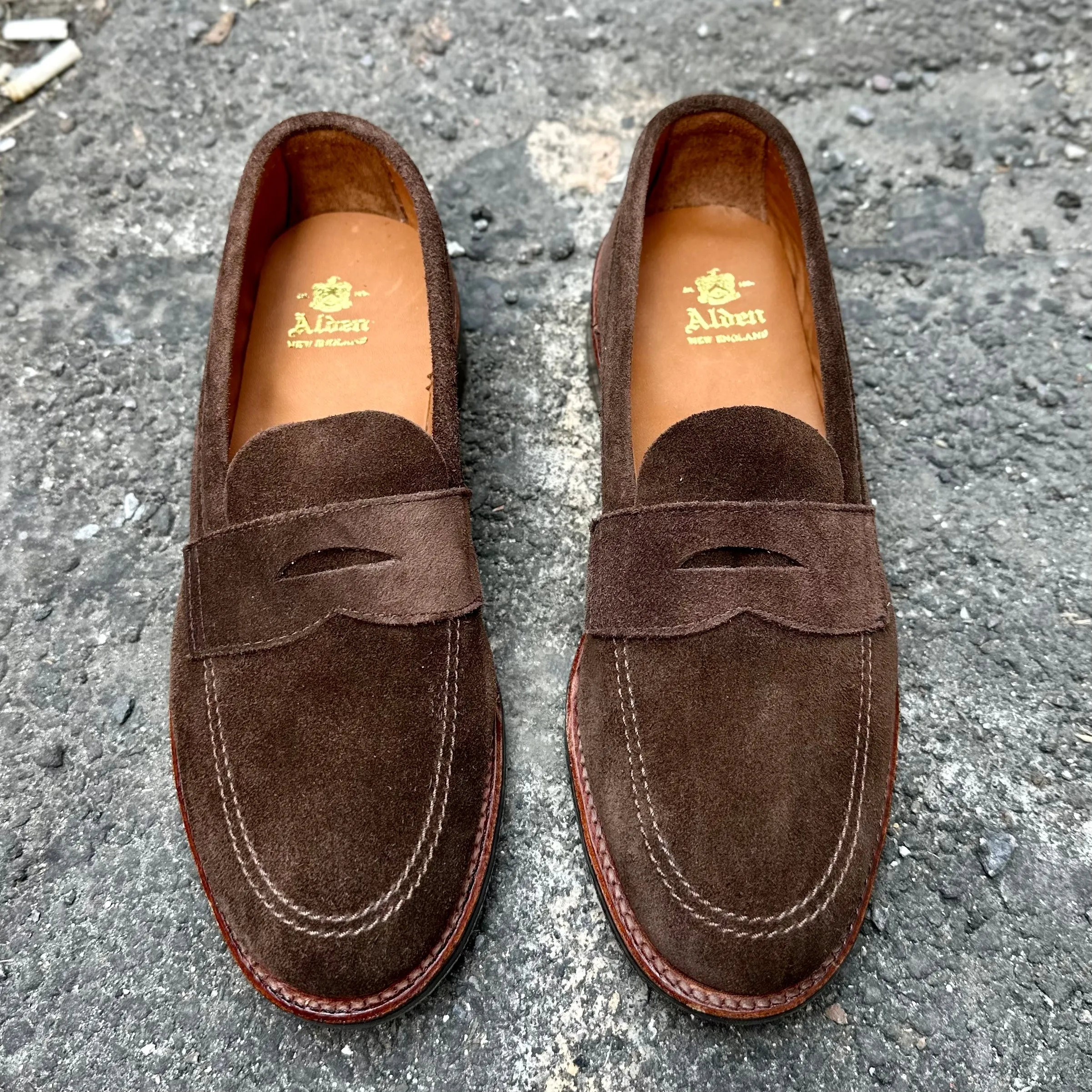 The 6224L Unlined Penny Mocc in Brown Suede by Alden Madison showcases a mocc toe design with stitching details on a rough gray surface. The insoles feature A. Albert in gold, paired with durable rubber soles for enhanced comfort and grip.