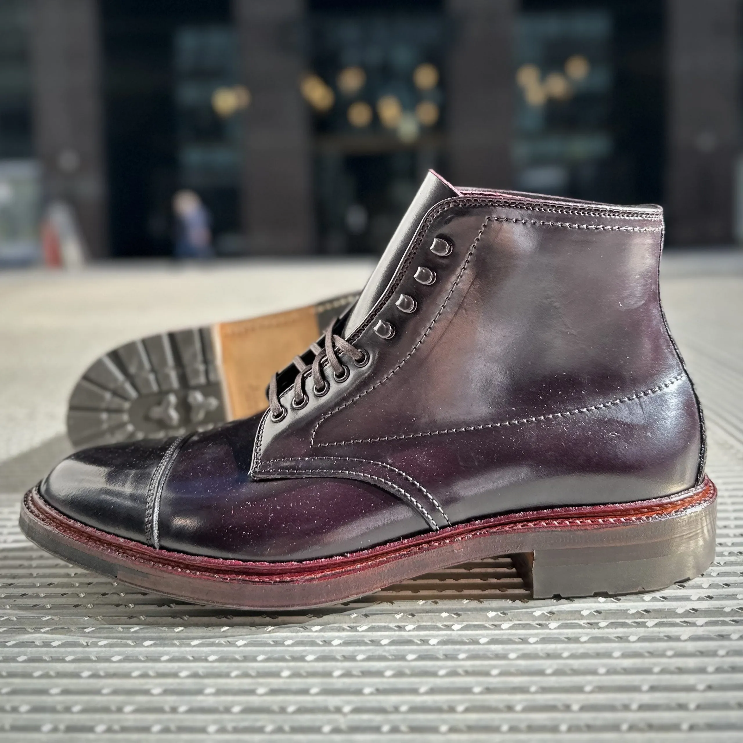 An Alden Madison D2824HC Cap Toe Boot in Color 8 Shell with a Commando Sole features red stitching and untied laces, resting on a textured surface with a blurred building faade in the background.