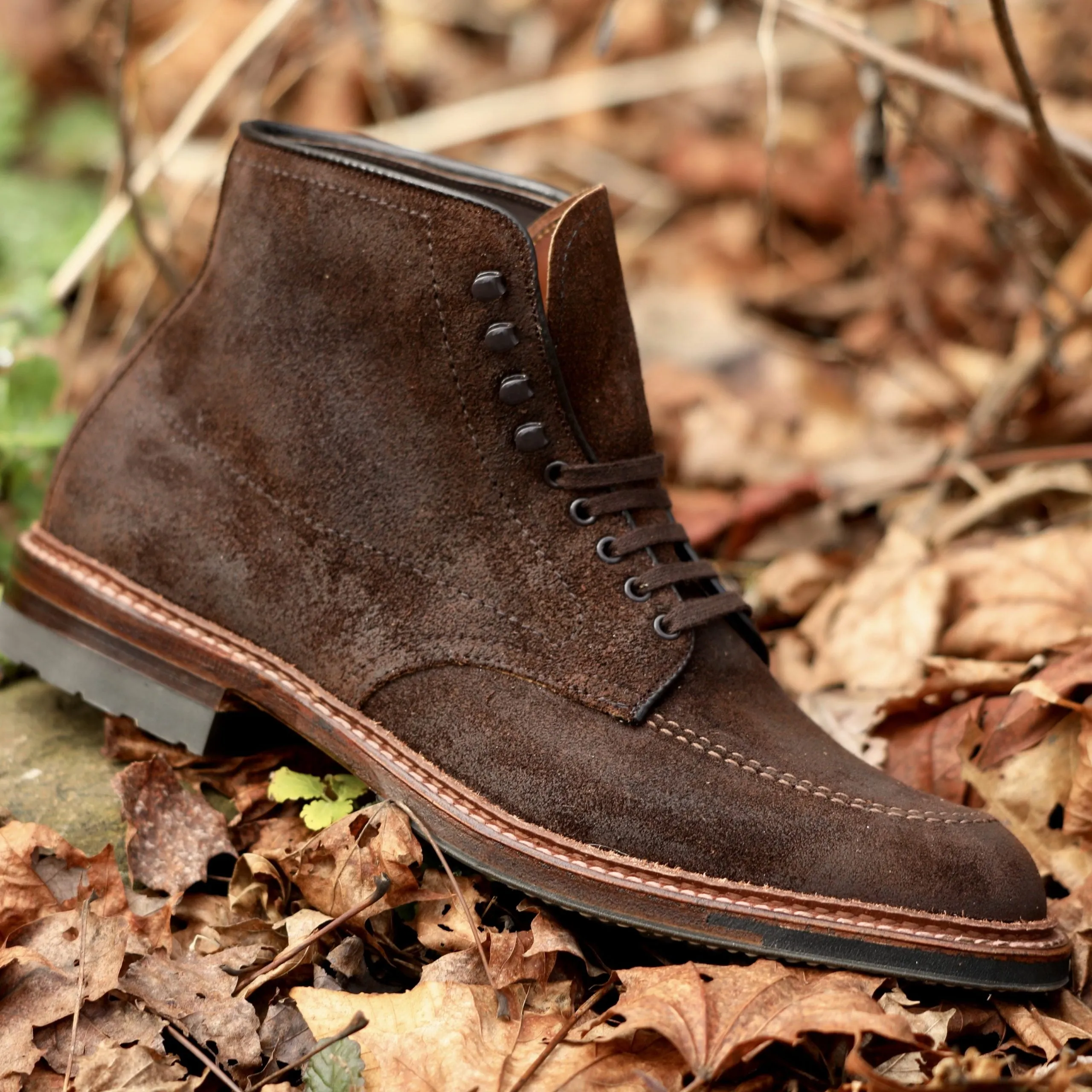 Close-up of the Alden Madison 4015HC - Indy Boot in Tobacco Reverse Chamois with a Commando Sole on dry autumn leaves. It highlights detailed stitching and metallic eyelets, showcasing craftsmanship and rugged design amidst brown leaves and green foliage.