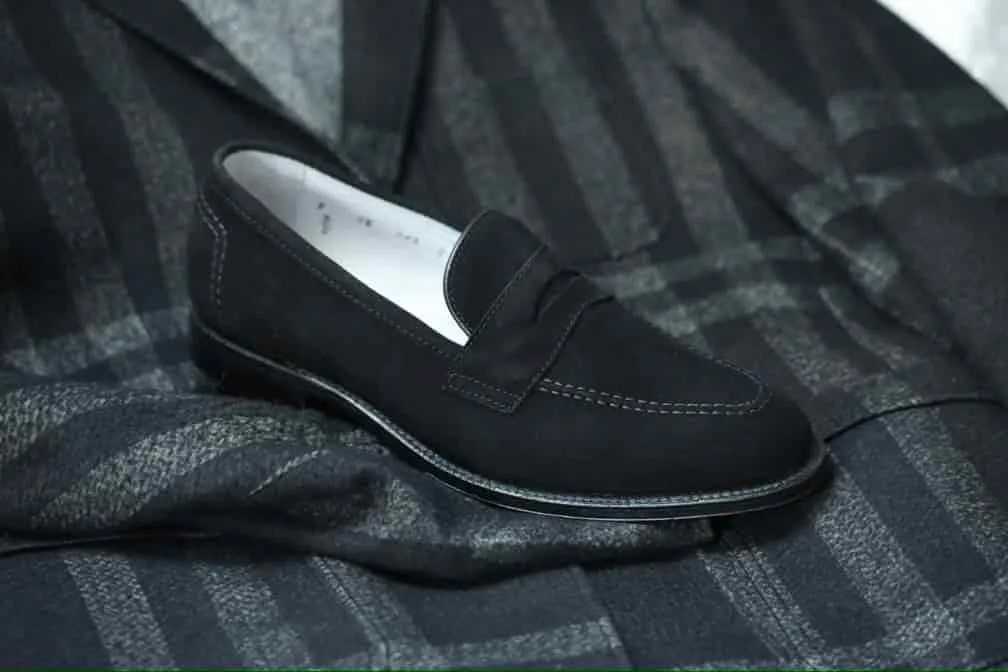 The D2229 - Madison Penny Loafer in Black Kid Suede by ALDEN SHOES sits on a dark plaid fabric, showcasing its sleek look and textured kid suede that contrasts with the smooth leather sole against the patterned background.