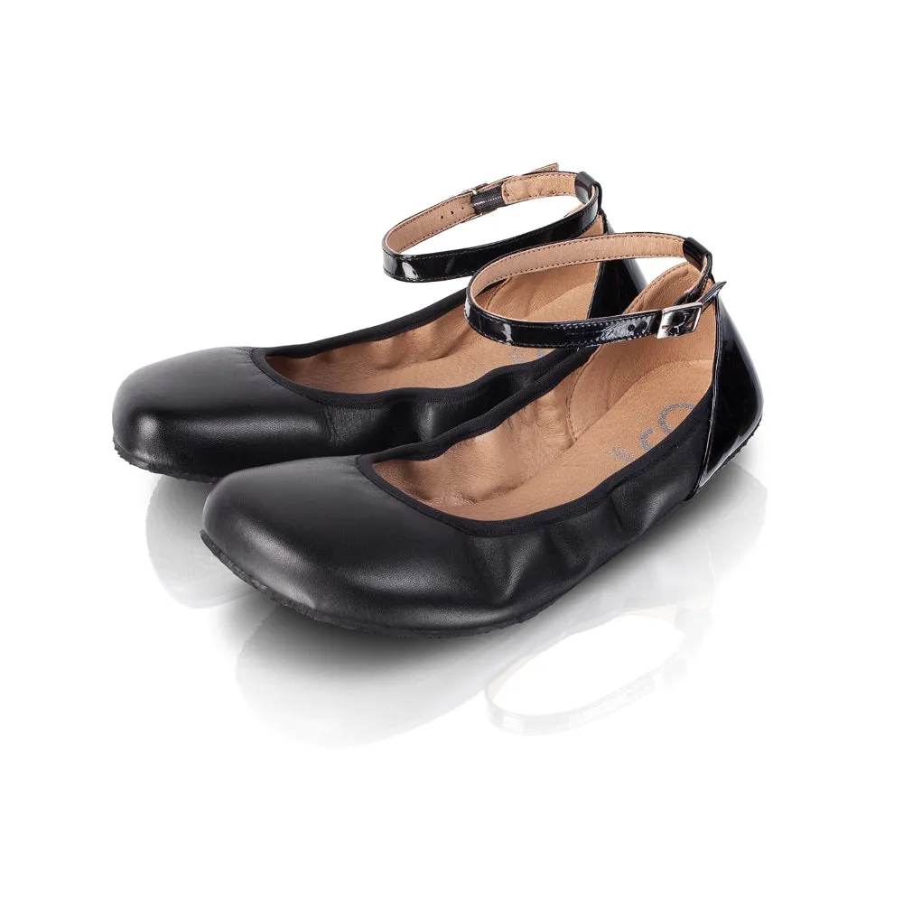 A photo of Shapen Tulip flats in black made of leather and rubber soles. Shoes have a gentle elastic around the perimeter of the opening, a patent leather heel cup, and an optional patent leather ankle strap. Both shoes are shown diagonally here from the f