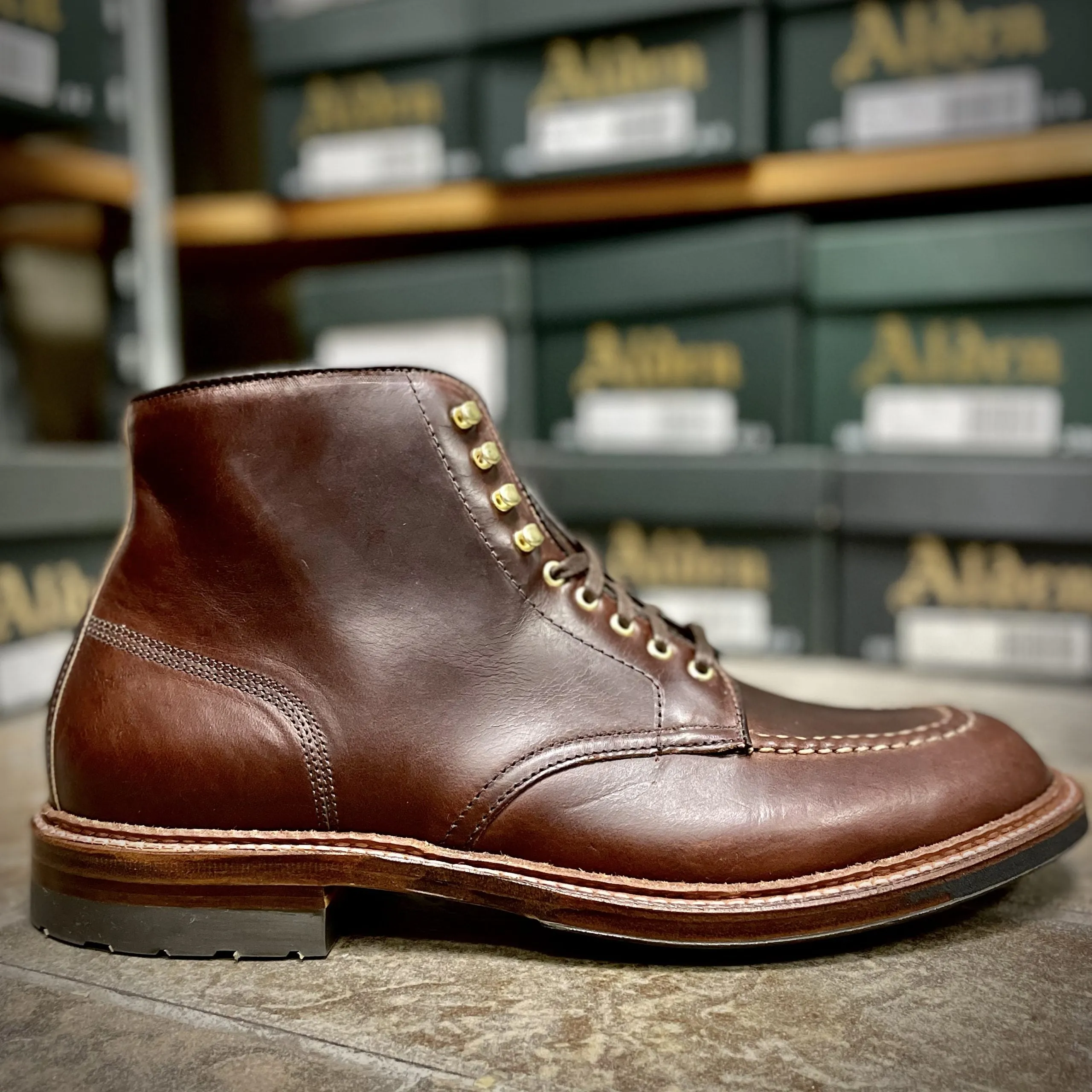 A close-up of Alden Madisons D1932HC - Dirty Bones Indy Boot in Brown Chromexcel, crafted with polished leather and featuring a stitched toe and gold eyelets, is showcased against a backdrop of neatly arranged green shoeboxes.