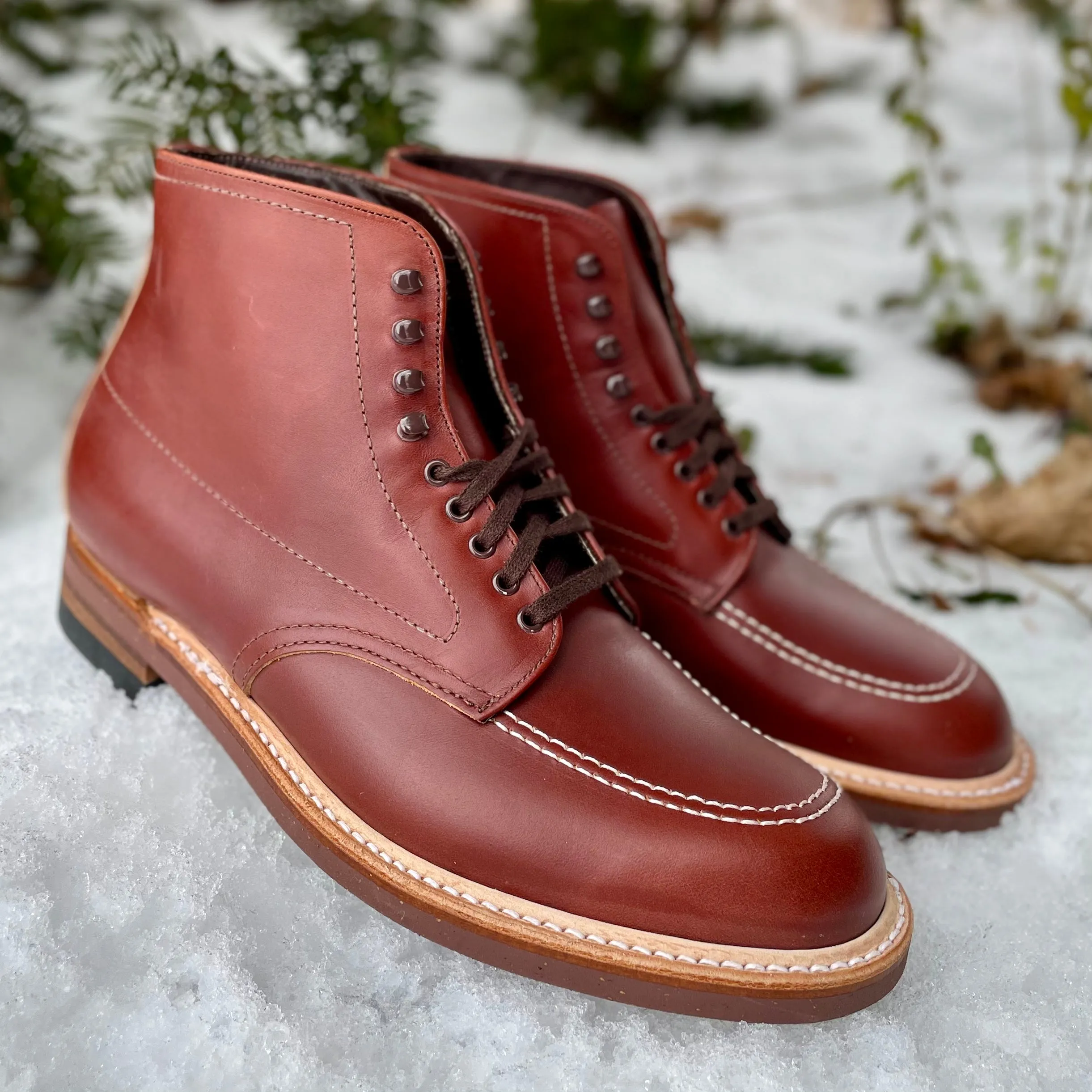 A pair of Alden Madison 405 - Indy Boot (Original) in Mid Brown Calfskin with black laces, featuring a moc toe design and contrasting stitching, set on snowy ground with green foliage in the background.