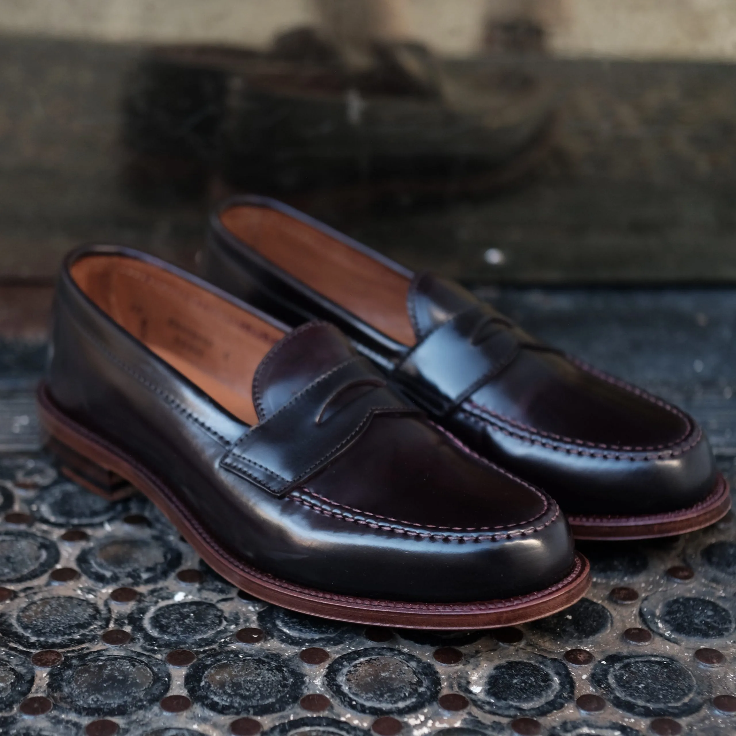 A pair of D6207 - Antique LHS in Color 8 Shell penny loafers from Alden Madison sits on a textured black rubber mat, highlighting their sleek design, leather sole, and light tan interior. The shoes feature intricate antique edging and detailed stitching, r