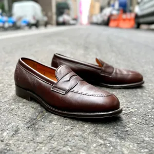 A pair of Alden Madisons 9694F Flex Penny Loafers in Brown Lady Calf, crafted on a Copley last with a polished finish, rests on a textured street surface, set against an urban backdrop with blurred elements like cars and barricades.