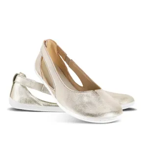 A photo of Be Lenka Bellissima flats with a leather upper and rubber soles. The flats are gold in color with a small stitching V detail in the front and a cut out design on the sides. Left shoe is shown from the right side with the right shoe propped up on