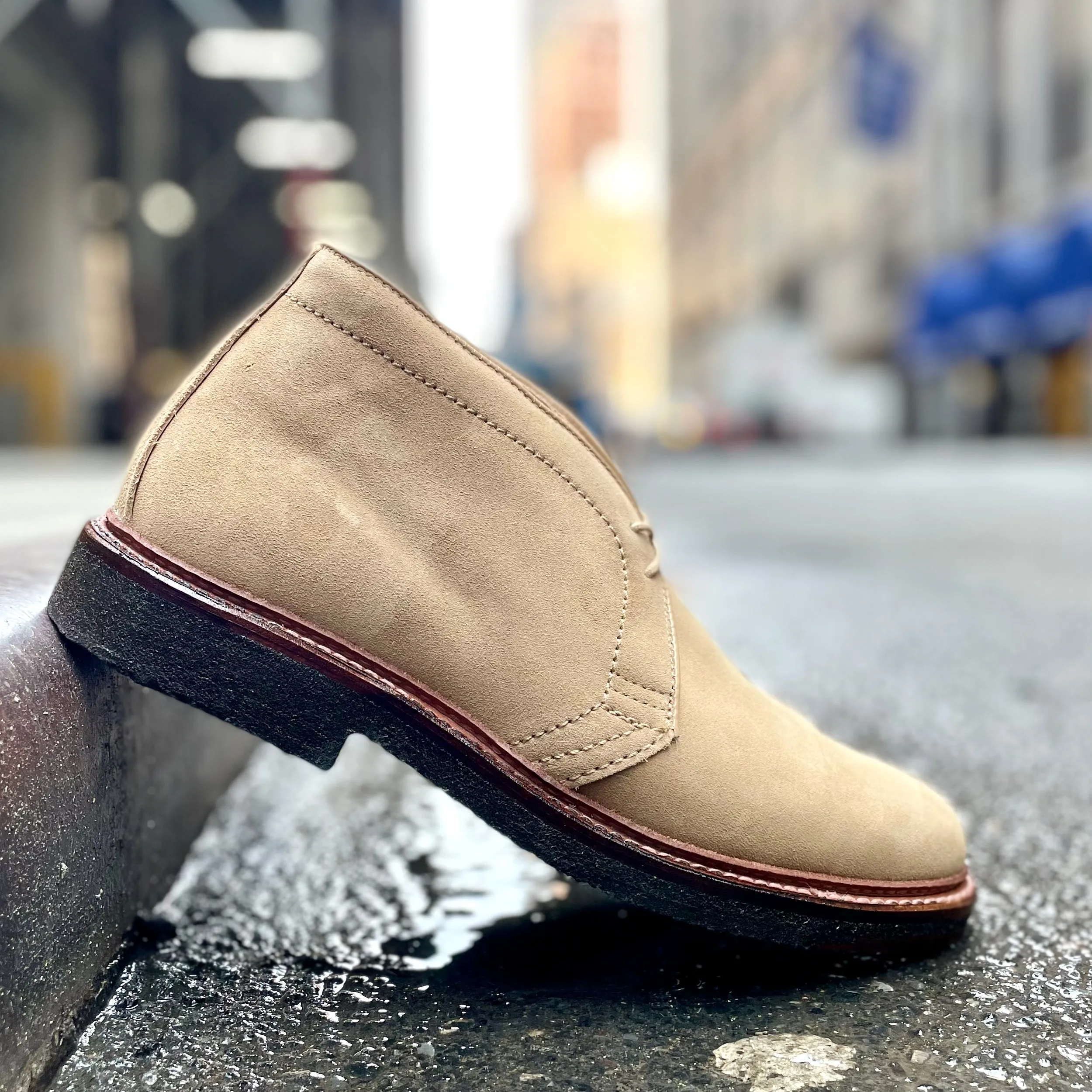The Alden Madison 14580 - Desert Chukka Boot in Tan Suede features a Barrie Last design with dark brown stitching and a crepe sole, resting on a curb against the blurred backdrop of a city street with towering buildings and soft lights.