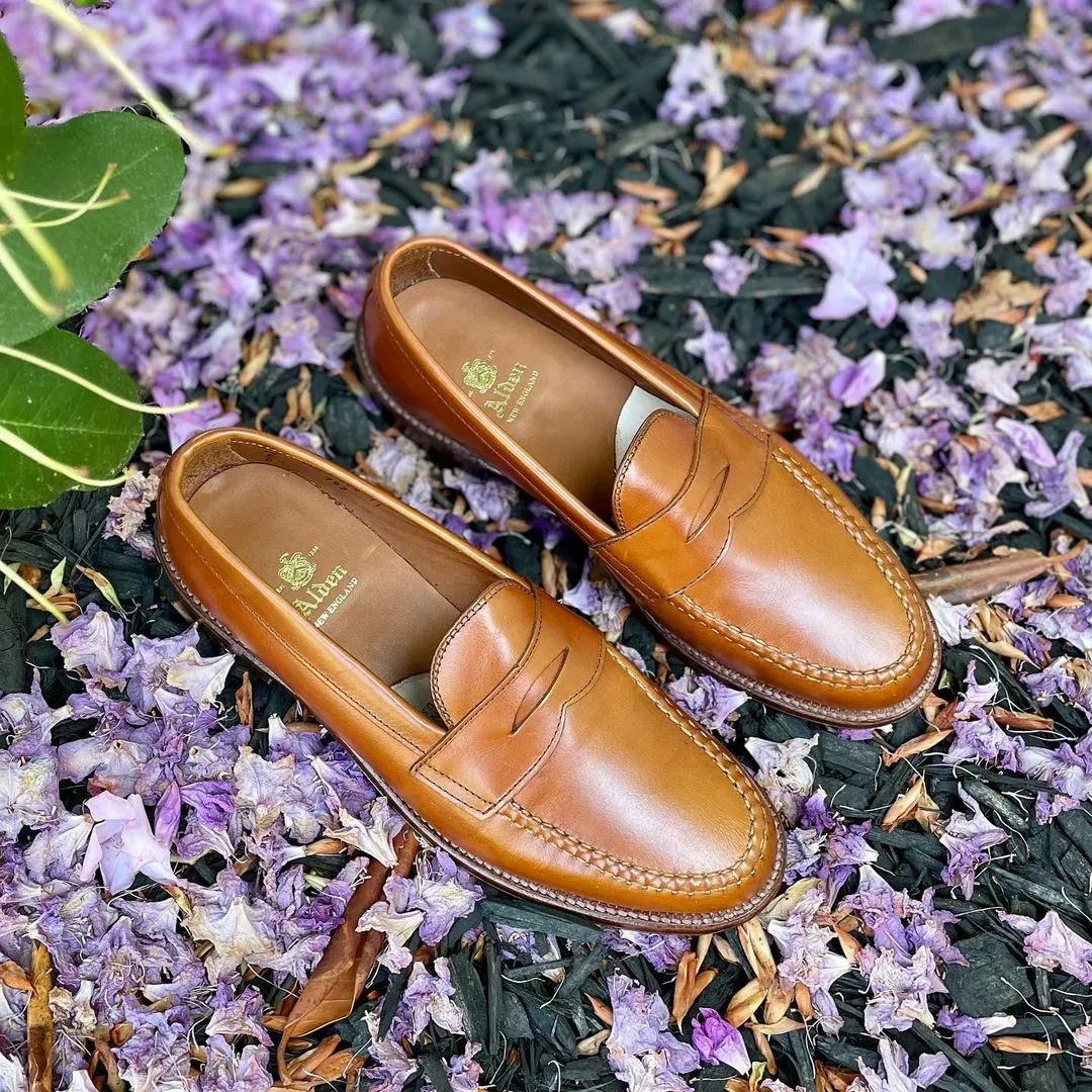 A pair of Alden Madisons 983 - Leisure Hand Sewn loafers in burnish tan calfskin rests on a purple flower and dark mulch. Featuring visible stitching and a branded logo on the insole, a large green leaf peeks from the top left corner to add a touch of natu