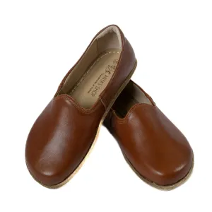 A photo of Brown Yasemin Leather loafers Designed by Anya with a leather upper and tan rubber soles. The loafers have a small curve up on the top of the foot for design. Shoe is shown with the left heel stacked on top of the right shoe opening against a wh