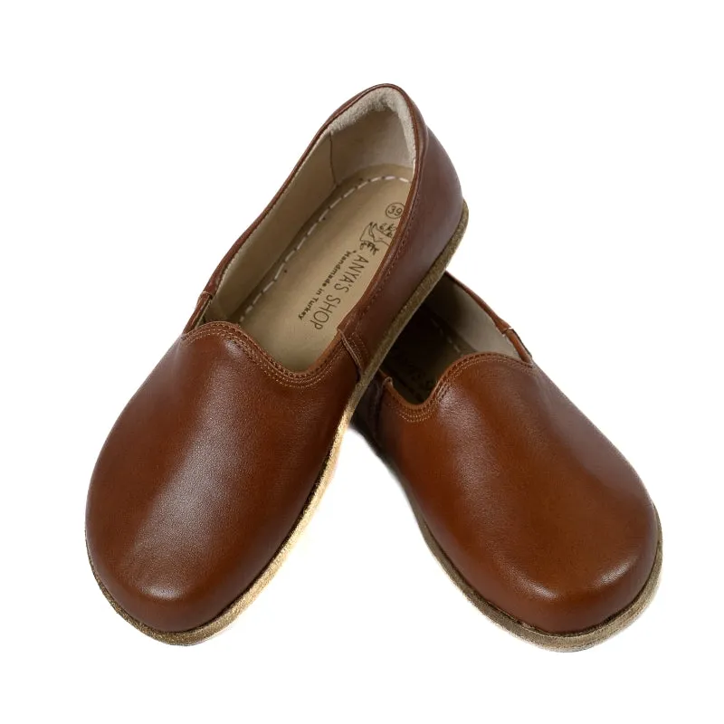 A photo of Brown Yasemin Leather loafers Designed by Anya with a leather upper and tan rubber soles. The loafers have a small curve up on the top of the foot for design. Shoe is shown with the left heel stacked on top of the right shoe opening against a wh
